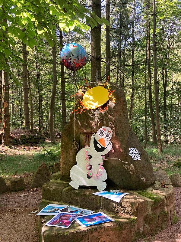 An einem Stein im Wald ist der Schneemann Olaf aus dem Film "Die Eiskönigin" aus Pappe angebracht. An dem Felsen hängt auch ein Luftballon mit dem Motiv und es liegen bunte Ausdrucke mit Disneyfiguren aus.