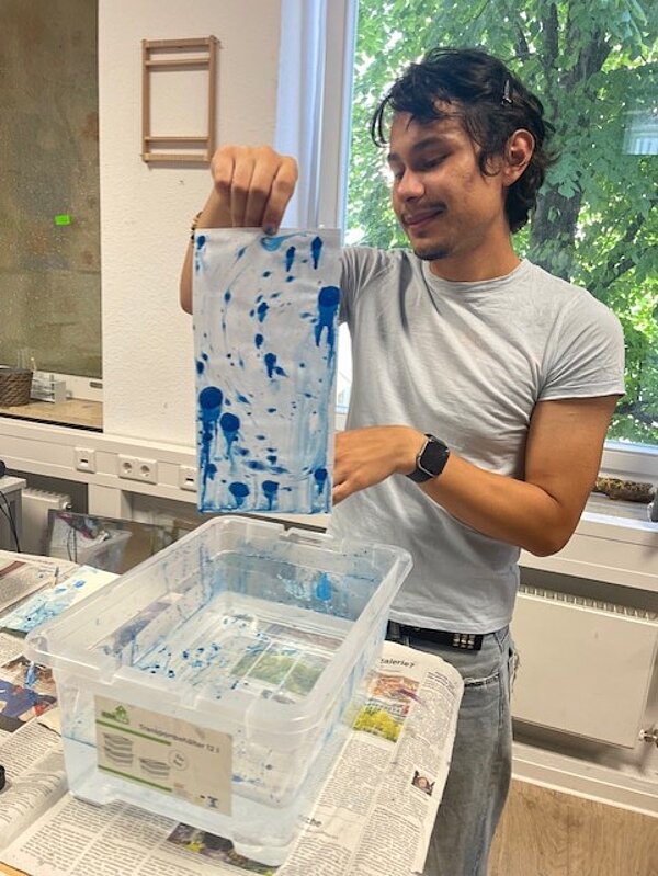 Eine Person mit grauem T-Shirt hält ein Blatt Papier mit blauen Farbmustern in die Höhe. Vor ihr steht eine durchsichtige Kunststoffwanne mit Wasser. Der Tisch ist mit Zeitungen bedeckt, und im Hintergrund sind Fenster zu erkennen.