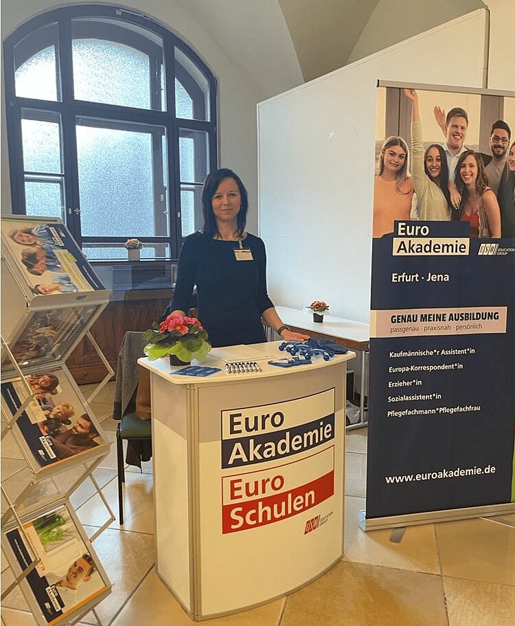 Erfolgreiche Messetermine Frau Gudernatsch steht hinter einem Stehtisch, auf den die Logos der Euro Akademie und Euro-Schulen gedruckt sind. Links neben hier ist ein Roll-up der Euro Akademien Erfurt und Jena aufgebaut, auf der die Ausbildungen beworben werden. Links neben ihr steht ein Ständer mit Broschüren zum Ausbildungsangebot.