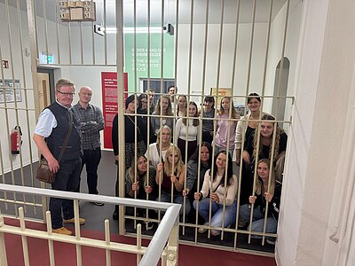Eine Gruppe Jugendlicher und zwei Begleitpersonen posiert hinter einem großen Metallgitter in einer historischen Ausstellung.