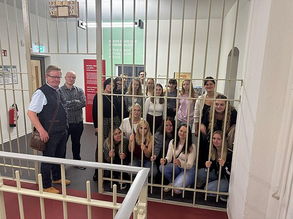 Eine Gruppe Jugendlicher und zwei Begleitpersonen posiert hinter einem großen Metallgitter in einer historischen Ausstellung.