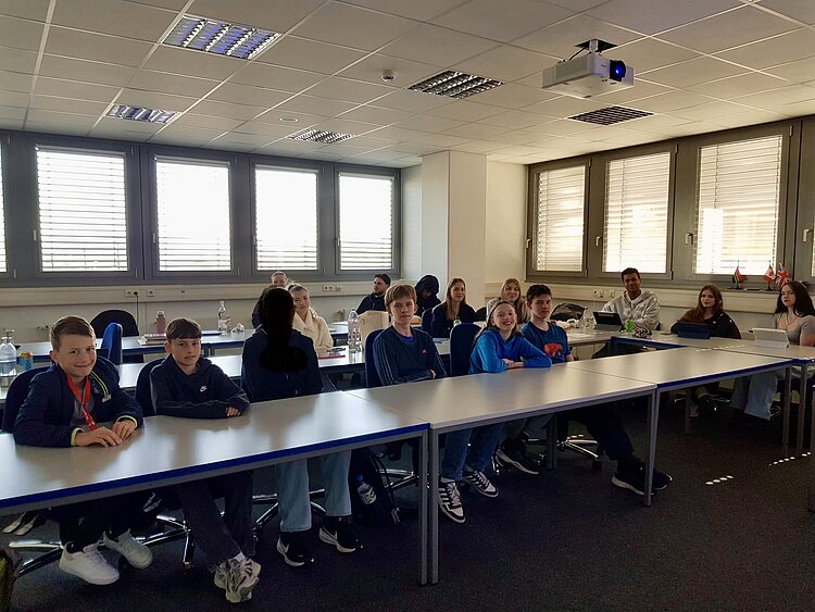 Gruppe von Jugendlichen sitzt in einem hellen Klassenraum an Tischen und schaut in die Kamera.