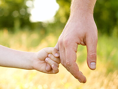 Eine Kinderhand hält den kleinen Finger einer erwachsenen Hand. Im Hintergrund ist grüne Natur unscharf zu erkennen.