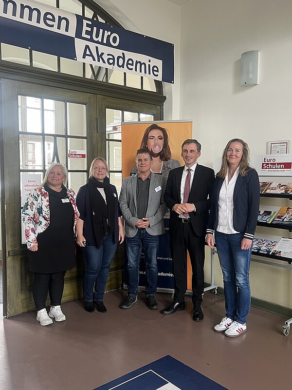 Sechs Personen stehen nebeneinander vor einer großen Glastür in einem Innenraum. Die Personen tragen Namensschilder und sind überwiegend formell oder halbformell gekleidet. Im Hintergrund hängt ein Schild mit der Aufschrift 'Euro Akademie' über der Tür. Rechts im Bild befindet sich ein Regal mit Broschüren und einem weiteren Schild mit der Aufschrift 'Euro Schulen'.
