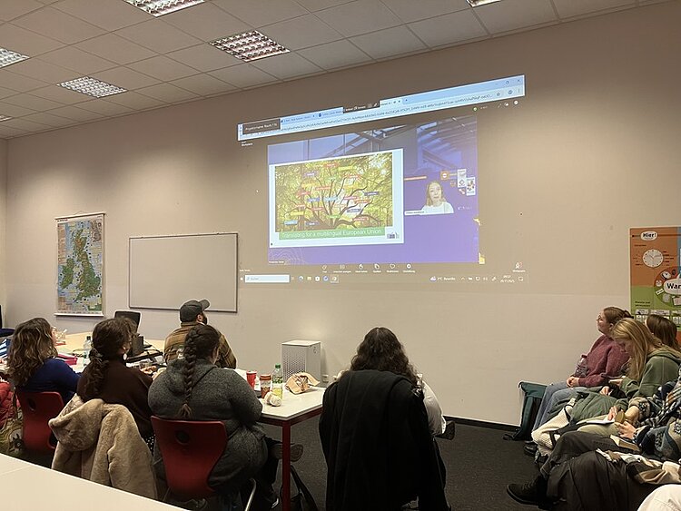 In einem hell erleuchteten Seminarraum sitzen mehrere Auszubildende der Euro Akademie Bamberg an einem langen Tisch und blicken auf eine Wandprojektion. Auf der Projektion ist eine Online-Veranstaltung der Generaldirektion Übersetzung (DGT) der Europäischen Kommission zu sehen, bei der ein farbiges Bild eines Baumes mit vielen Ästen und Blättern gezeigt wird. Rechts neben dem Bild ist ein kleines Videofenster mit einer Person zu sehen, die an der Online-Veranstaltung teilnimmt. Im Raum sind an der linken Wand eine Landkarte und an der rechten Wand ein orangefarbenes Plakat mit Text und Symbolen angebracht. Die Auszubildenden tragen unterschiedliche Kleidung, einige mit Jacken und Schals, und sitzen auf roten und weißen Stühlen. Auf dem Tisch liegen verschiedene Gegenstände wie Getränke und Schreibmaterialien. Die Atmosphäre wirkt konzentriert und lernorientiert.