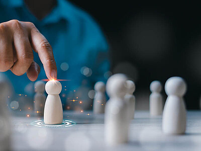 Große weiße Spielfiguren stehen auf einem Tisch. Eine Person tippt mit der Fingerspitze auf eine Figur und dabei leuchtet der Berührungspunkt auf. Um diese Spielfigur leuchten mehrere Lichtpunkte auf und sie steht in einem digital erzeugten Kreis.