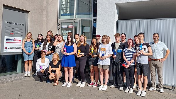 Erster Schultag: Alle an die Zuckertüten In einem Innenhof steht eine Gruppe junger Mädchen und Jungen in sommerlicher Kleidung, hinter ihnen ist ein geschlossenes Tor und eine offene Tür, die in ein Treppenhaus führt. Die jungen Menschen halten kleine Schultüten/Zuckertüten in den Händen. Einige ältere Personen, offenbar Lehrerinnen und Lehrer, haben keine solchen Tüten in der Hand.