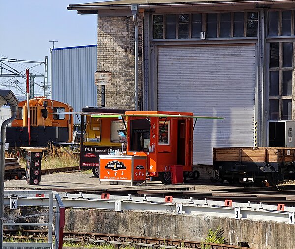 Ein orangefarbener Imbisswagen mit der Aufschrift 'Drink Truck' steht auf einem Bahnhofsgelände. Im Hintergrund ist ein historischer Zug zu sehen, und zwei Männer stehen vor dem Wagen und eine Frau steht an der Bedientheke. Ein großes Rolltor und ein Schild mit der Aufschrift 'Haus für Dampflok' sind ebenfalls sichtbar.