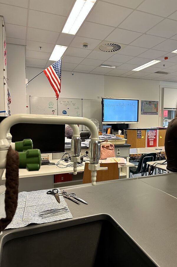 Ein Klassenzimmer mit einer US-amerikanischen Flagge an der Wand. Im Vordergrund sind ein Wasserhahn zu sehen und auf einem Handtuch liegen Scheren und Pinzetten. Im Hintergrund sind ein Whiteboard und ein Bildschirm mit einer Präsentation zu sehen.