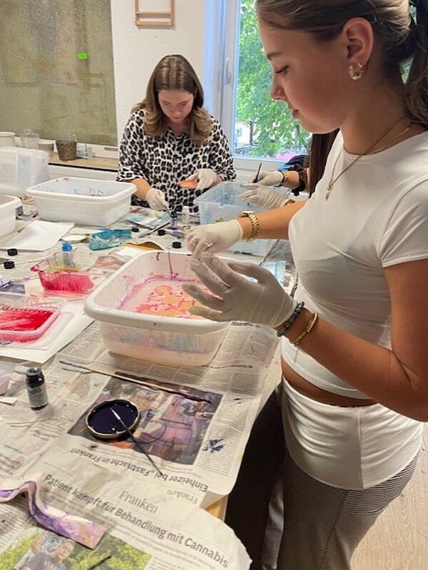 Zwei Personen arbeiten konzentriert an einem Tisch. Die Person im Vordergrund, mit weißem T-Shirt und Handschuhen, bearbeitet eine weiße Kunststoffwanne mit farbigen Mustern. Im Hintergrund sitzt eine weitere Person mit gemustertem Oberteil und arbeitet ebenfalls an einem ähnlichen Projekt. Der Tisch ist mit Zeitungen ausgelegt, und es sind verschiedene Materialien wie Farbdosen und Pinsel zu sehen.