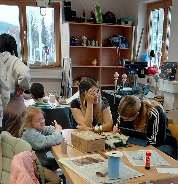 In einem Bastelraum sitzen mehrere Kinder und junge Erwachsene an einem Tisch, arbeiten an Kartonmodellen und reden miteinander; im Hintergrund stehen Regale mit Materialien und Werkzeugen.