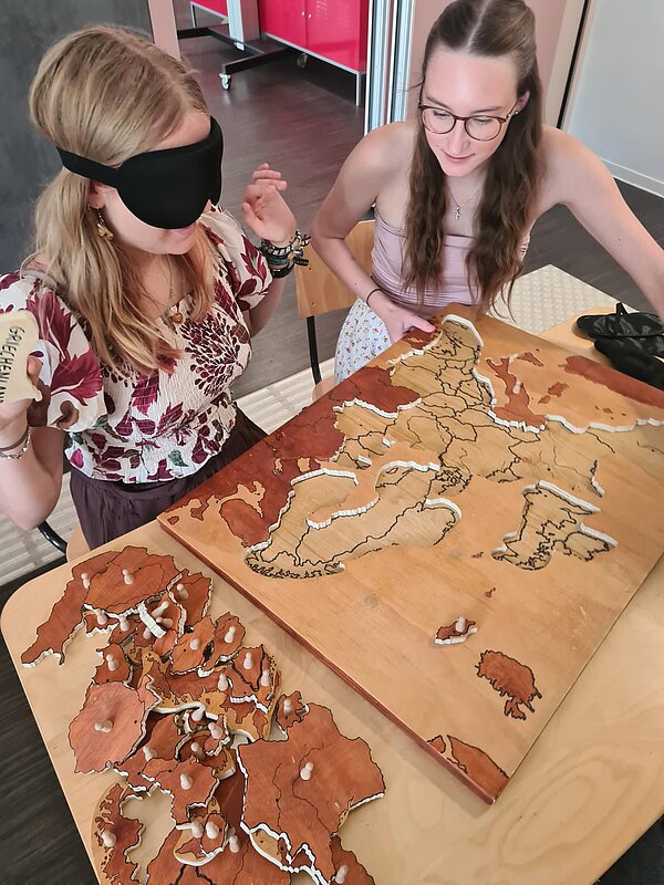 Zwei Schülerinnen sitzen an einem Tisch, eine von ihnen trägt eine Augenmaske, die andere eine Brille. Vor ihnen auf dem Tisch liegt eine Weltkarte aus Holz. Die Länder sind, wie in einem Puzzle, ausgestanzt und lassen sich an einem kleinen Knubel in der Mitte des jeweiligen Landes hochheben und an der passenden Stelle der Weltkarte einsetzen.