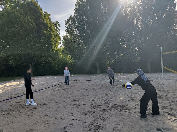 Vier Personen spielen Beachvolleyball auf einem Sandplatz. Im Hintergrund sind hohe Bäume zu erkennen. Ein Sonnenstrahl fällt durch die Bäume auf das Spielfeld.