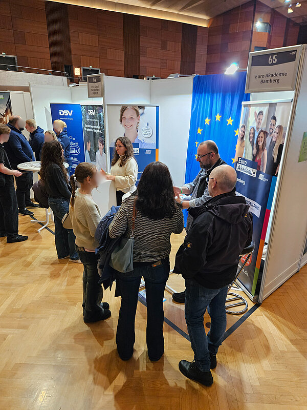 Mehrere Besucherinnen und Besucher stehen in einer Messehalle vor dem Stand der Euro Akademie Bamberg und führen Beratungsgespräche mit den Auszubildenden und Lehrkräften.