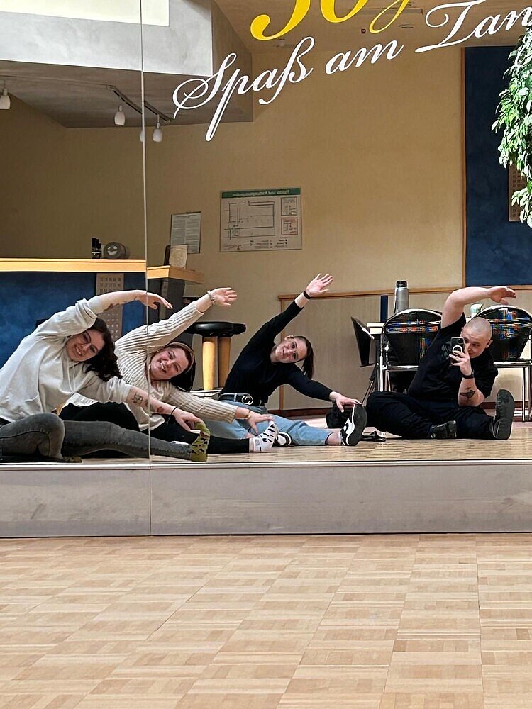 Fit für die Prüfung mit Bewegung Ein Selfie im Spiegel: Die vier Personen auf dem Bild schauen in die Spiegelwand im Tanzsaal und verrenken sich dabei, indem sie im Sitzen mit ihrer linken Hand an ihre linke Fußspitze greifen. Der einzige Mann auf dem Bild hält dabei sein Smartphone in der anderen Hand.