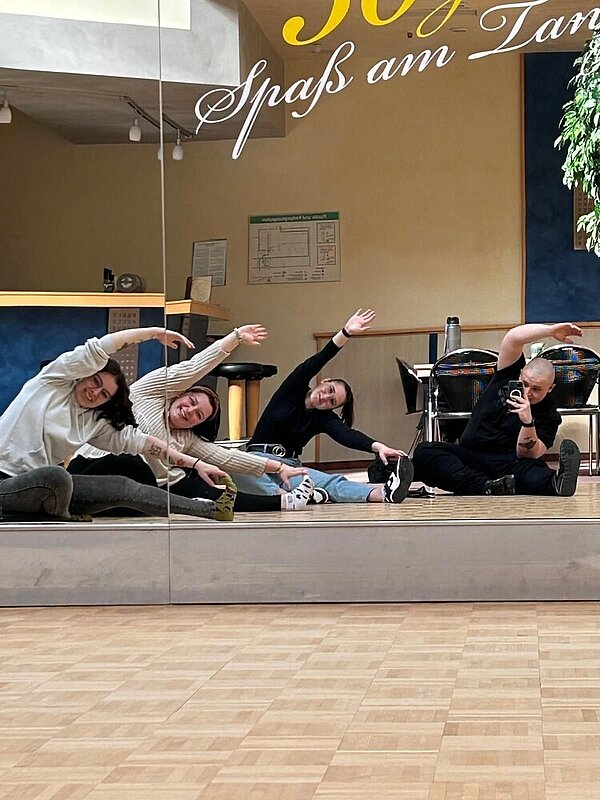 Fit für die Prüfung mit Bewegung Ein Selfie im Spiegel: Die vier Personen auf dem Bild schauen in die Spiegelwand im Tanzsaal und verrenken sich dabei, indem sie im Sitzen mit ihrer linken Hand an ihre linke Fußspitze greifen. Der einzige Mann auf dem Bild hält dabei sein Smartphone in der anderen Hand.