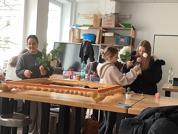 Drei Schüler*innen binden Kunstblumen zu kleinen Bouquets. Auf dem Tisch liegt umgedreht der Bilderrahmen.