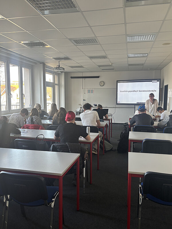 In einem hellen Klassenzimmer lauschen Jugendliche einem Vortrag; auf einer Leinwand ist eine Präsentation zu sehen.