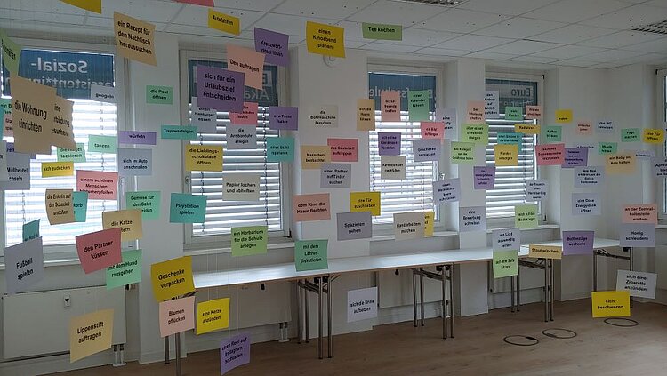 Ein Klassenzimmer oder Seminarraum, an dessen Wänden und Fenstern zahlreiche bunte Zettel mit verschiedenen schriftlichen Aufgaben oder Szenarien für Rollenspiele oder Übungen befestigt sind. Die Zettel enthalten kurze Anweisungen oder Situationsbeschreibungen auf Deutsch, wie z.B. 'ein Rezept für den Nachbarshund ausleihen' oder 'Tee kochen'. Im Raum stehen mehrere Tische und Stühle, die für Gruppenarbeit vorbereitet sind.