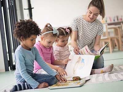 Drei Kinder hocken mit ihrer Erzieherin in der Kita auf dem Boden und schauen Bilderbücher an.
