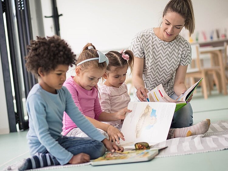 Drei Kinder hocken mit ihrer Erzieherin in der Kita auf dem Boden und schauen Bilderbücher an.
