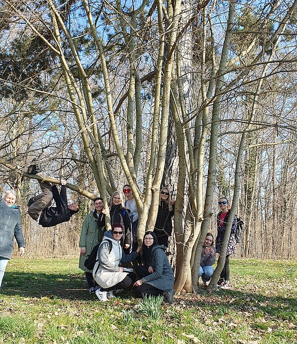 Exkursion in die Natur Eine Gruppe posiert fröhlich um einen Baum im Wald. Eine Person hängt kopfüber von einem Ast, andere stehen lachend drumherum. Die Stimmung ist ausgelassen und entspannt.