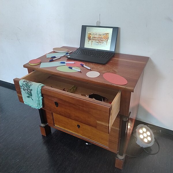 Eine offene kleine Holzkommode, auf der ein geöffneter Laptop steht und Kugelschreiber und Zettel auf ihr liegen. 