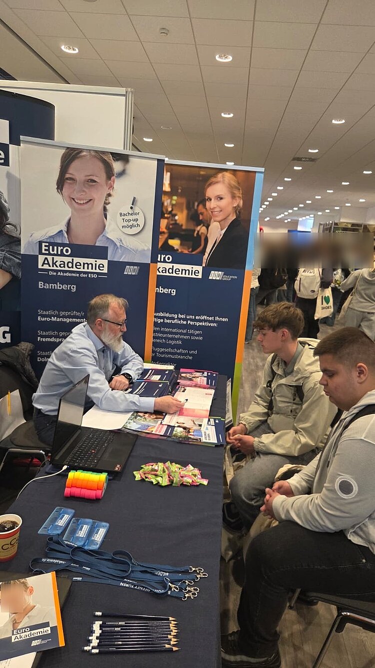 Ein Mitarbeiter der Euro Akademie berät zwei Besucher an einem Messestand mit Flyern, Give-aways und Roll-up-Bannern.
