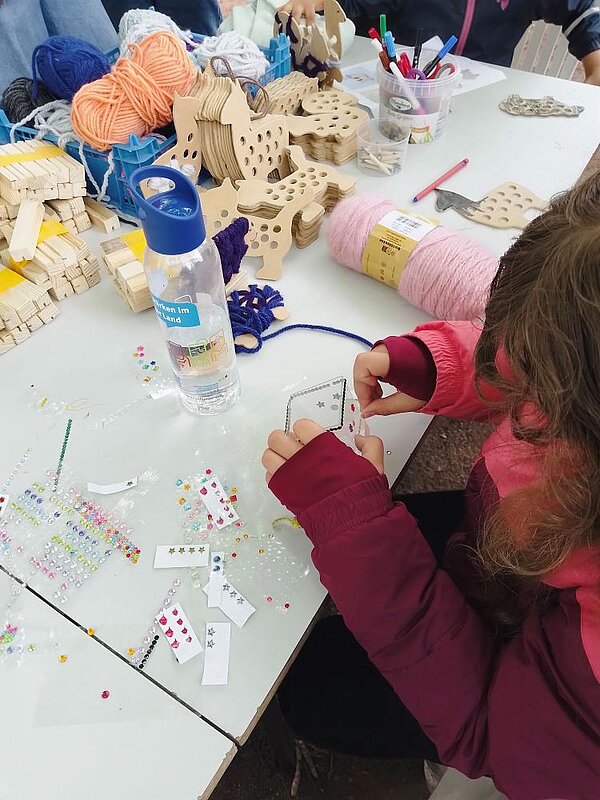 Ein Kind in einer pink-roten Jacke bastelt an einem Tisch mit Wolle, Holzfiguren und anderen Materialien. Im Vordergrund steht eine Wasserflasche.