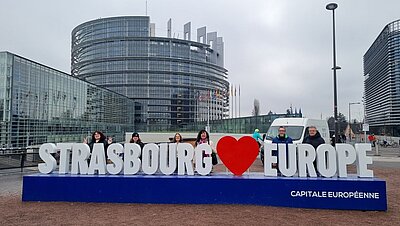 Gruppe von sechs Personen posiert lächelnd vor dem Europäischen Parlament in Straßburg. Die Personen stehen hinter einem großen blauen Schild mit der weißen Aufschrift ‚STRASBOURG ♥ EUROPE‘ und dem Zusatz ‚CAPITALE EUROPÉENNE‘. Im Hintergrund ist das moderne, runde Glasgebäude des Europäischen Parlaments mit vielen Flaggenmasten zu sehen. Der Himmel ist bewölkt.