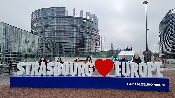 Gruppe von sechs Personen posiert lächelnd vor dem Europäischen Parlament in Straßburg. Die Personen stehen hinter einem großen blauen Schild mit der weißen Aufschrift ‚STRASBOURG ♥ EUROPE‘ und dem Zusatz ‚CAPITALE EUROPÉENNE‘. Im Hintergrund ist das moderne, runde Glasgebäude des Europäischen Parlaments mit vielen Flaggenmasten zu sehen. Der Himmel ist bewölkt.