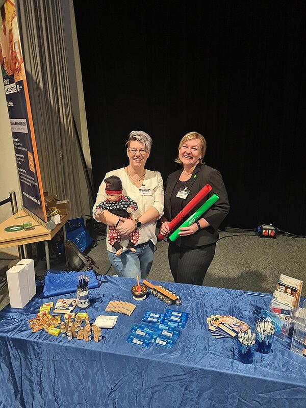 Zwei Mitarbeiterinnen posieren hinter dem Messestand lächelnd für ein Foto.