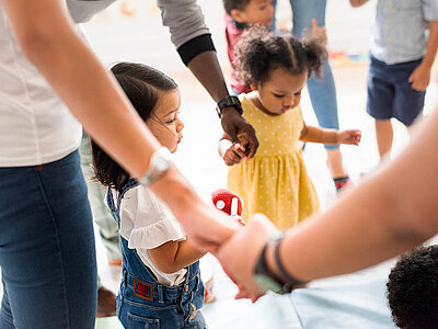 Eine Gruppe von Kindern und Erwachsenen steht im Kreis und hält sich an den Händen. Ein Kind in einem gelben Kleid steht in der Mitte und hält einen roten Ball in der Hand. Die Szene wirkt fröhlich und spielerisch.