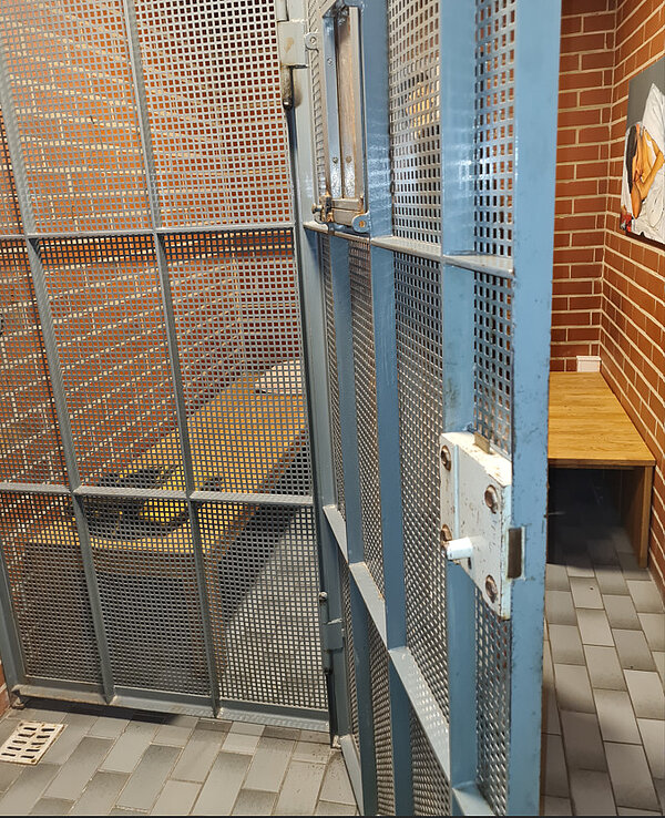Blick in eine kleine Zelle mit Metalltür und Gitter. Dahinter steht eine Holzbank.