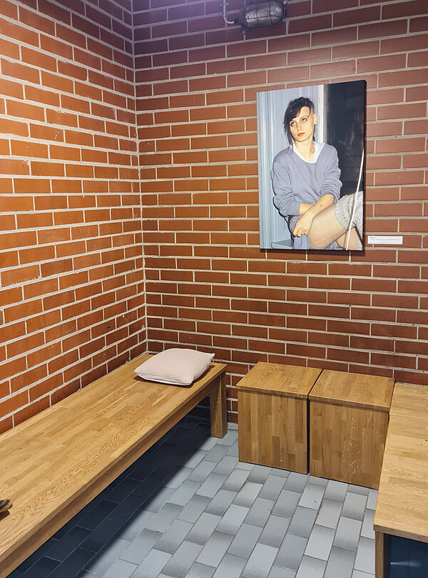 Kleine Zelle mit roten Backsteinwänden, Holzbank und einem Bild einer jungen Frau an der Wand.