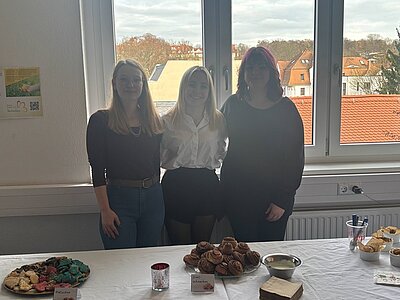Drei weibliche Auszubildende stehen hinter einem Tisch, auf dem verschiedene Backwaren arrangiert sind. Die linke Frau trägt ein schwarzes Oberteil und blaue Jeans, die mittlere Frau ein weißes Hemd und einen schwarzen Rock, die rechte Frau ein schwarzes Oberteil. Der Tisch ist mit mehreren Tellern und Schalen gedeckt, auf denen unter anderem Zimtschnecken und andere kleine Gebäckstücke liegen. Im Hintergrund sind große Fenster mit Blick auf Dächer und Bäume zu sehen. Die Szene spielt im Rahmen des Tages der offenen Tür an der Euro Akademie Zwickau.