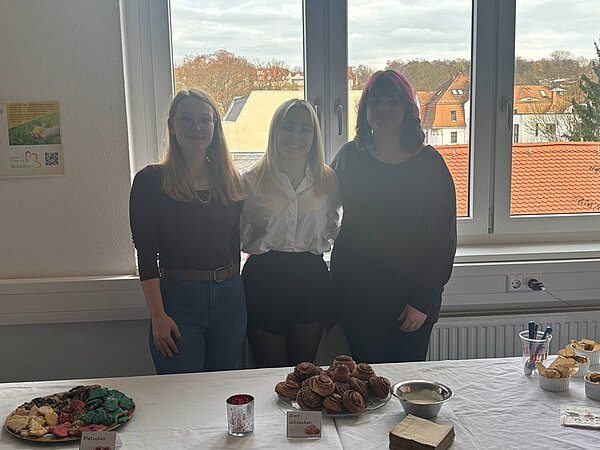 Drei weibliche Auszubildende stehen hinter einem Tisch, auf dem verschiedene Backwaren arrangiert sind. Die linke Frau trägt ein schwarzes Oberteil und blaue Jeans, die mittlere Frau ein weißes Hemd und einen schwarzen Rock, die rechte Frau ein schwarzes Oberteil. Der Tisch ist mit mehreren Tellern und Schalen gedeckt, auf denen unter anderem Zimtschnecken und andere kleine Gebäckstücke liegen. Im Hintergrund sind große Fenster mit Blick auf Dächer und Bäume zu sehen. Die Szene spielt im Rahmen des Tages der offenen Tür an der Euro Akademie Zwickau.