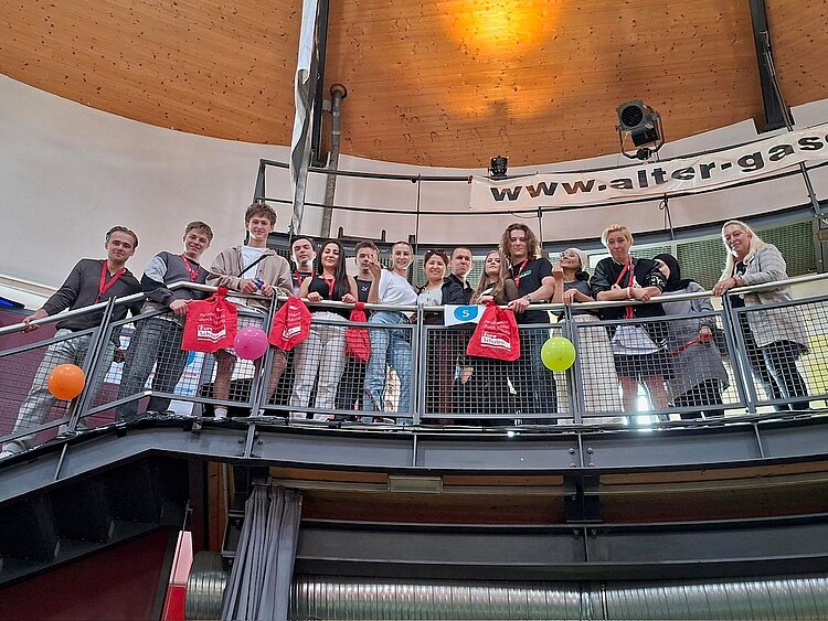 Eine Gruppe von etwa 14 Personen steht auf einer umlaufenden Galerie in einem Veranstaltungsgebäude mit Holzkuppeldecke. Am Geländer hängen bunte Luftballons und rote Taschen. Die Aufnahme wurde von unten nach oben fotografiert.