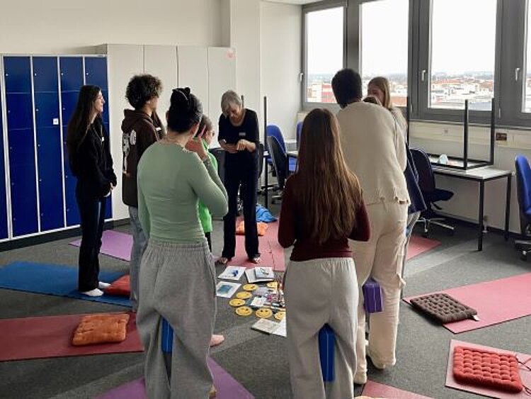 Mehrere Personen stehen um eine Sammlung von Karten und Gegenständen auf dem Boden. Sie unterhalten sich und scheinen sich über ihre Erfahrungen auszutauschen. Die Yogamatten und Kissen liegen noch im Hintergrund, und eine ältere Frau moderiert die Unterhaltung.