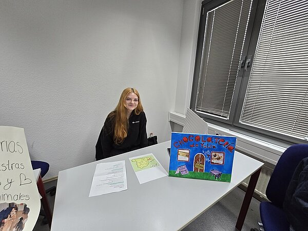 Eine Auszubildende mit langen, hellen Haaren sitzt an einem Tisch in einem schlicht eingerichteten Raum. Auf dem Tisch liegen mehrere Blätter Papier, darunter ein handgezeichneter Plan und ein beschriftetes Blatt. Vor der Person steht ein farbig gestaltetes Plakat mit Fotos und dem Titel in großen Buchstaben. Im Hintergrund sind ein Fenster mit Jalousien und eine weiße Wand zu sehen. Links am Bildrand steht ein weiteres Plakat mit handgeschriebenem Text.
