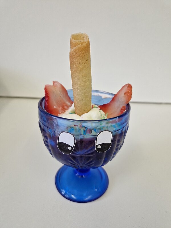 Gesund und gruselig: Kitaessen zu Halloween Blauer Eisbecher, mit einer Kugel Vanilleeis, links und rechts eine Scheibe Erdbeere und in der Mitte ein aufrecht eingestecktes Waffelröllchen.