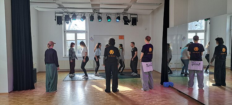 Mehrere Teilnehmende stehen in einem hellen Bewegungsraum mit Spiegelwand und Bühnenbeleuchtung und üben gemeinsam eine Szene oder Choreografie.