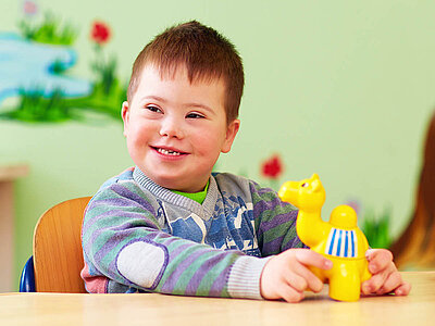 Ein lächelndes Kind mit Down-Syndrom sitzt an einem Tisch und spielt mit einem gelben Spielzeugpferd. Das Kind trägt einen gestreiften Pullover und wirkt glücklich und engagiert.