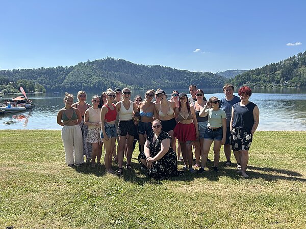 Gruppenbild mit jungen lächelnden Menschen vor einem See und bewaldeten Hügeln; links ist ein Bootsanleger zu sehen.