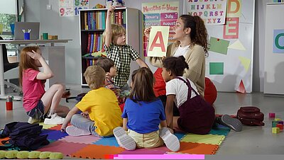 Mehrere Kinder sitzen auf einem Noppenteppich in bunten Farben und spielen zusammen mit einer weiblichen Betreuungskraft. Der Raum ist bunt und mit viel Spielzeug eingerichtet.