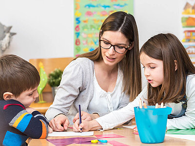 Eine Frau mit langen, dunklen Haaren und Brille sitzt mit zwei Kindern an einem Tisch. Sie trägt ein hellgraues Oberteil und hilft den Kindern beim Malen oder Schreiben, wobei sie einen Stift in der Hand hält. Neben ihr sitzen ein Junge in einem langärmeligen Shirt mit blauen, orangefarbenen und grauen Streifen sowie ein Mädchen in einem hellblauen Oberteil mit dunkler Schürze. Auf dem Tisch liegen bunte Papierstücke, Stifte und ein blauer Utensilienbehälter. Im Hintergrund sind bunte Poster und eine Pflanze zu sehen. Die Atmosphäre wirkt konzentriert, fördernd und lernorientiert.