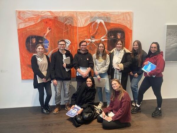 Eine Gruppe von neun Personen, die vor einem großen, abstrakten Kunstwerk in einer Galerie posieren. Einige stehen, andere sitzen auf dem Boden. Mehrere Personen halten Notizbücher oder Bücher in den Händen.