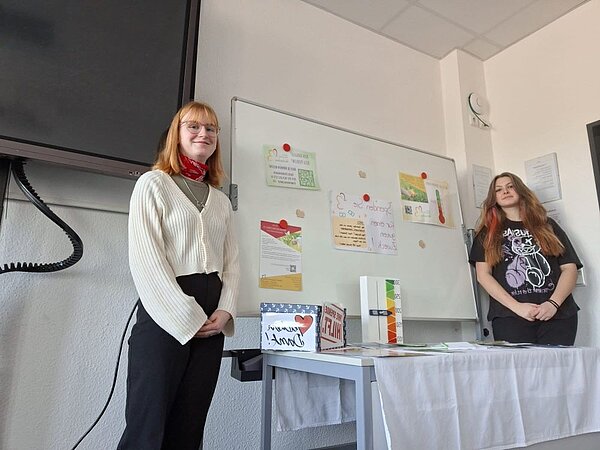 Zwei junge Frauen stehen vor einer Pinnwand in einem Klassenzimmer. Die Pinnwand ist mit verschiedenen Postern, Notizen und einem Thermometer-Poster dekoriert. Auf einem Tisch vor der Pinnwand liegen Informationsmaterialien.