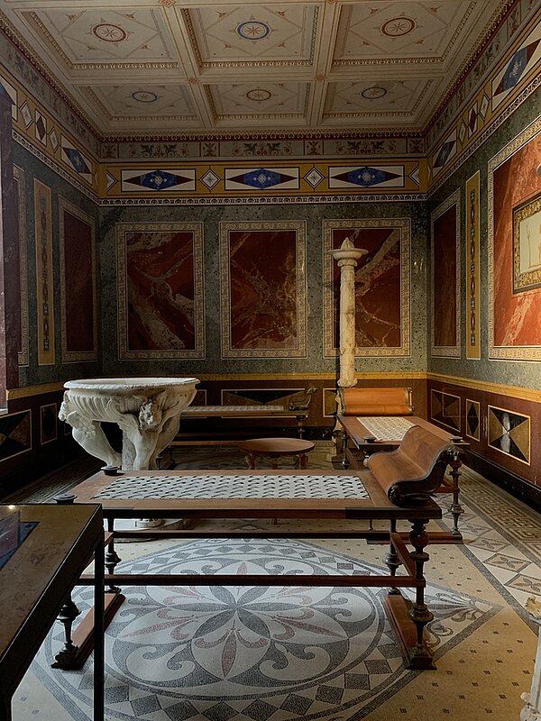 Eintauchen in die römische Kultur Ein dunkler Raum des Pompejanums ist ausgestattet mit einem altertümlichen Wasserbecken und verschiedenen Liegen. Die Fliesen auf dem Boden, an der Wand und an der Decke haben unterschiedliche Muster und Farben.