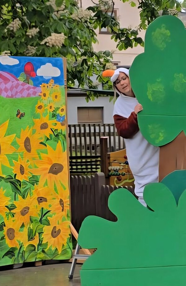 Bunte bemalte Kulisse mit großen Sonnenblumen, Schmetterlingen und einer Burg auf einem Hügel. Daneben steht eine Person in einem Tierkostüm, die teilweise von einer großen, grün bemalten Pappfigur in Blattform verdeckt wird. Im Hintergrund sind Bäume und ein Holzzaun zu sehen.